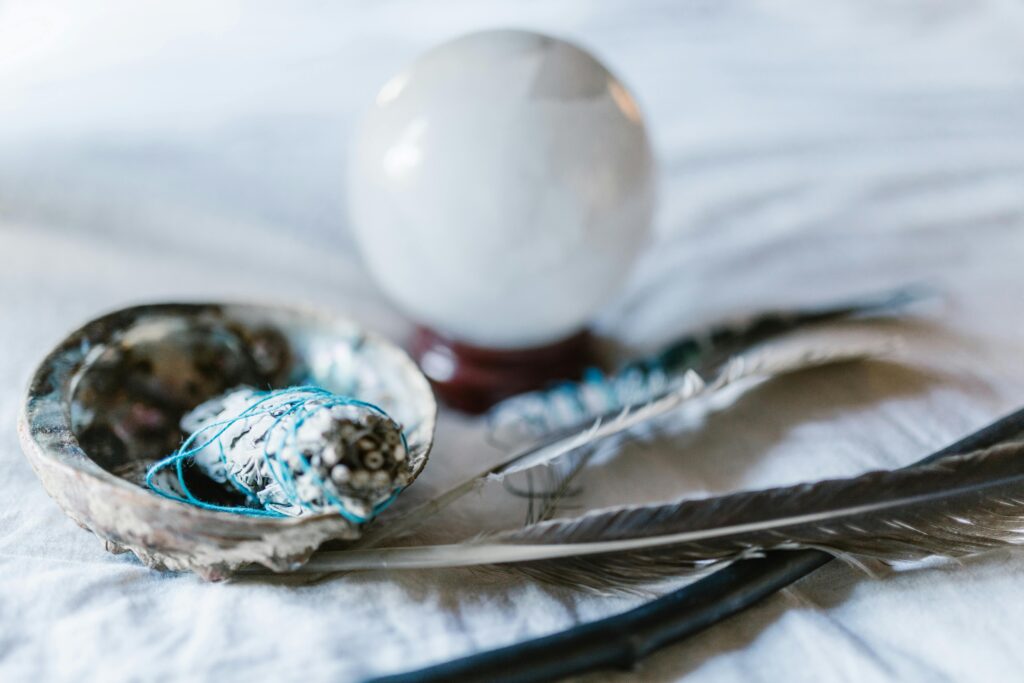 A white crystal sphere placed behind a sage smudge stick in an abalone shell with feathers used for cleansing rituals.