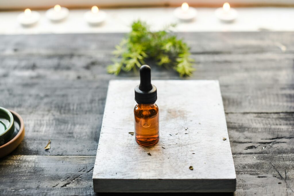 Small amber glass bottle with a dropper placed on a wooden surface, surrounded by candles and herbs in a peaceful setting.