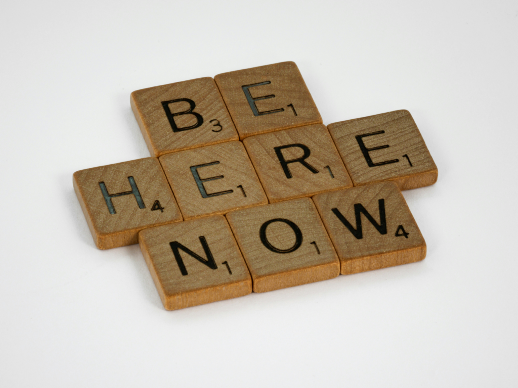 Wooden letter tiles spelling the word βhere now,β symbolising presence and awareness