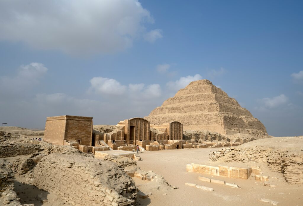 Ancient stepped pyramid and surrounding stone ruins under a clear blue sky, showcasing an archaeological site in a desert landscape.