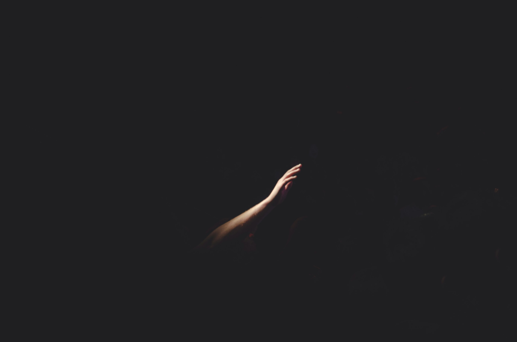 A single hand reaching through darkness toward light, symbolizing observing the darkness without judgment and allowing awareness to gently illuminate inner conflict.