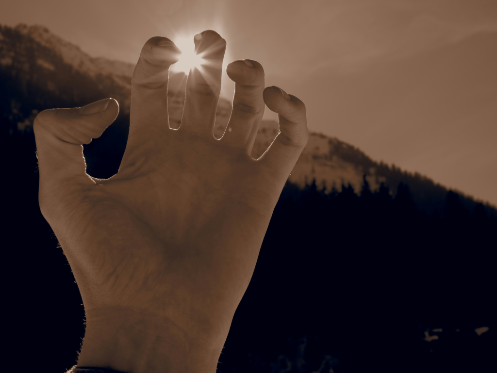 A human hand reaching toward sunlight breaking through a mountainous landscape, symbolising awareness, acceptance, and the gentle meeting of light within darkness.